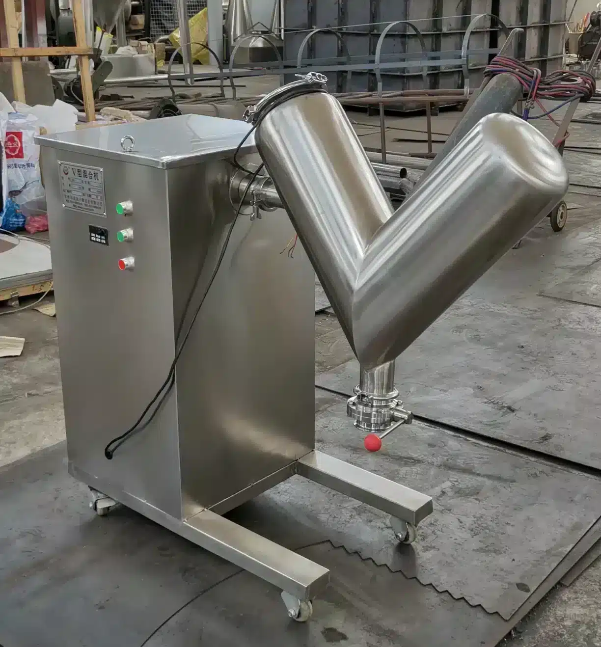 The image shows a large stainless steel V-shaped blender or mixer inside what appears to be an industrial manufacturing facility or workshop. The V blender has two cylindrical mixing chambers joined in a V configuration and is mounted on a movable stainless steel frame or stand. Various pipes, hoses and other equipment can be seen in the background of the cluttered workspace. V型混合机