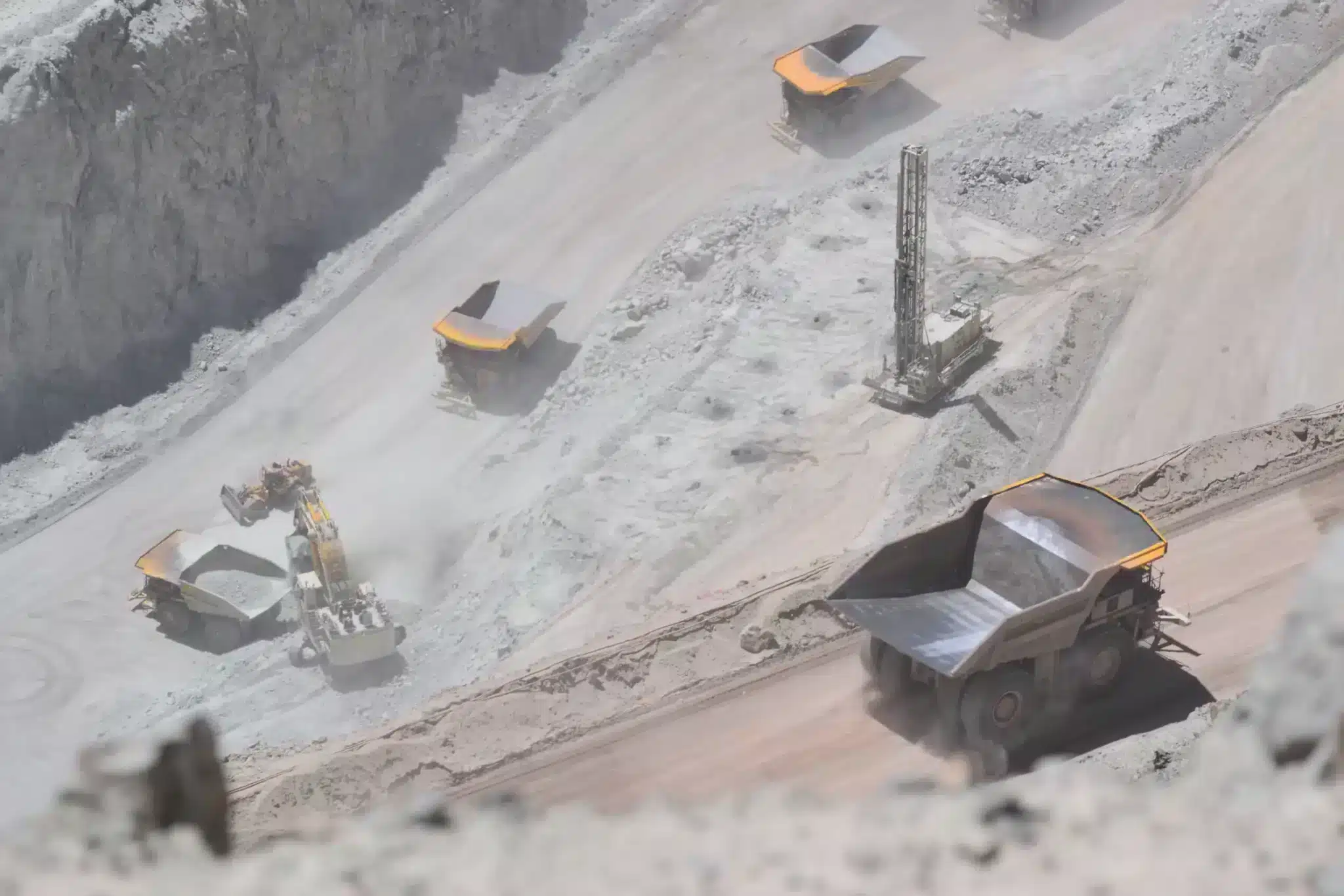 An active open-pit mining operation with large machinery at work. Several heavy dump trucks with yellow beds are visible transporting materials, while an excavator loads one of them. A drilling rig stands in the background amidst a landscape of exposed earth and rock.