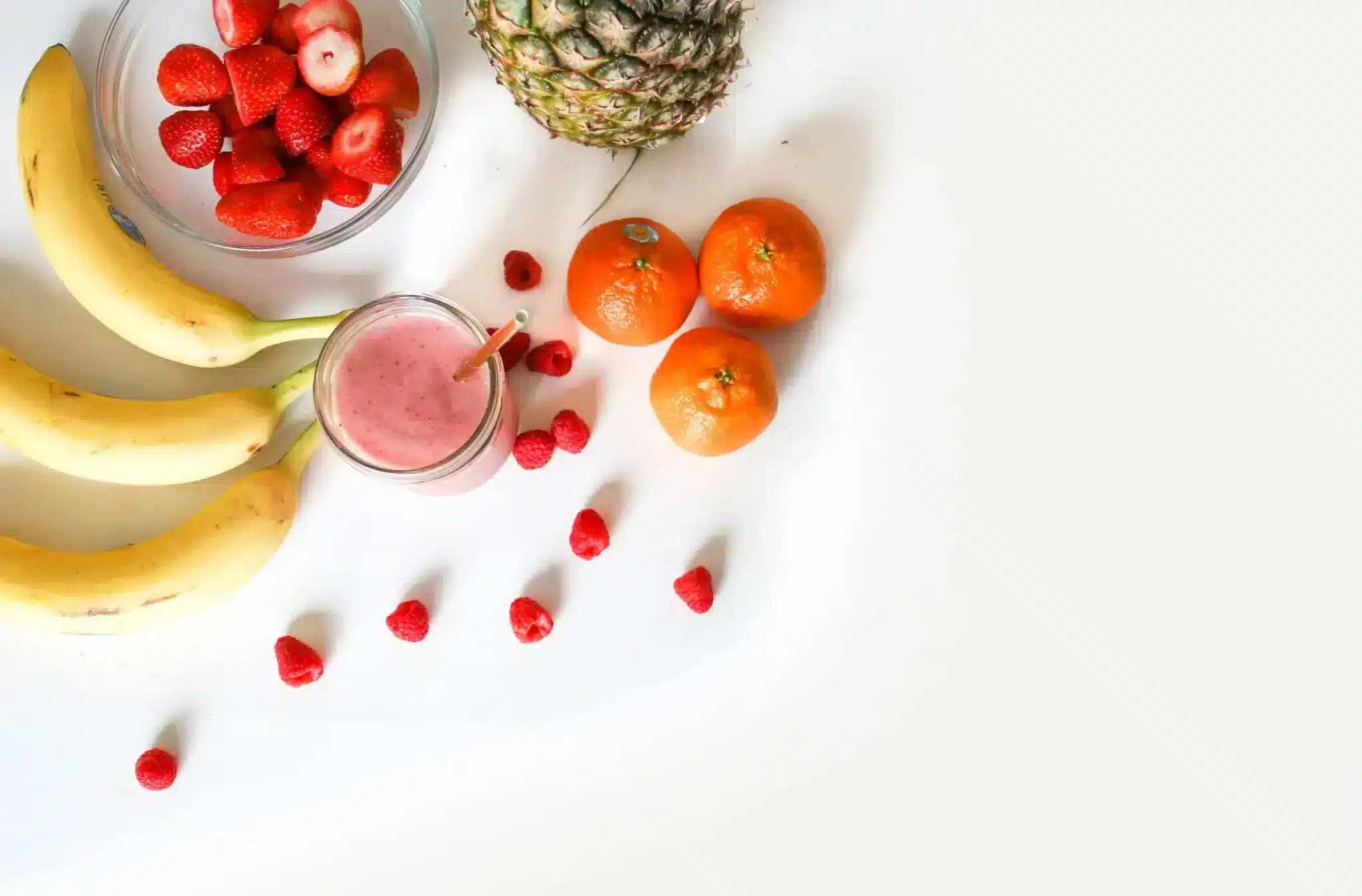 A bright and healthy arrangement of fruits on a white surface, featuring ripe bananas, a pineapple, several mandarins, a bowl of strawberries, scattered raspberries, and a glass of pink smoothie with a straw.