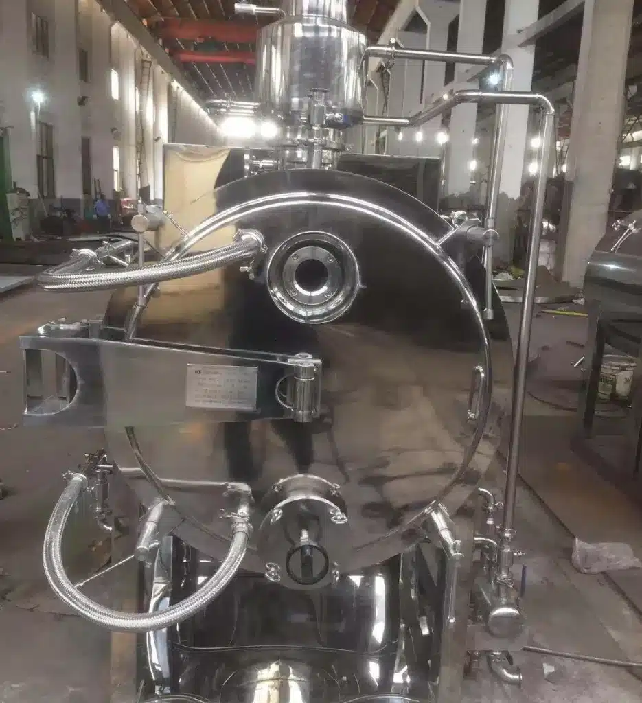 A Rake Dryer in an industrial setting, with a polished stainless steel exterior, visible piping, and a front opening for processing, set against an indoor backdrop.