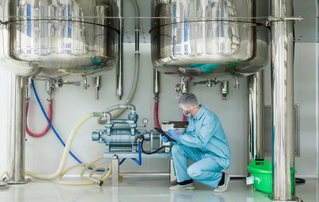 A technician in blue protective clothing and hairnet is squatting while examining a clipboard and a digital device in a clean industrial environment. Above him, two large, shiny stainless steel vessels are suspended from the ceiling, connected to various pipes and hoses in blue, pink, and white. The machinery appears to be part of a sophisticated processing system, possibly for chemicals or pharmaceuticals, with a focus on hygiene and precision.