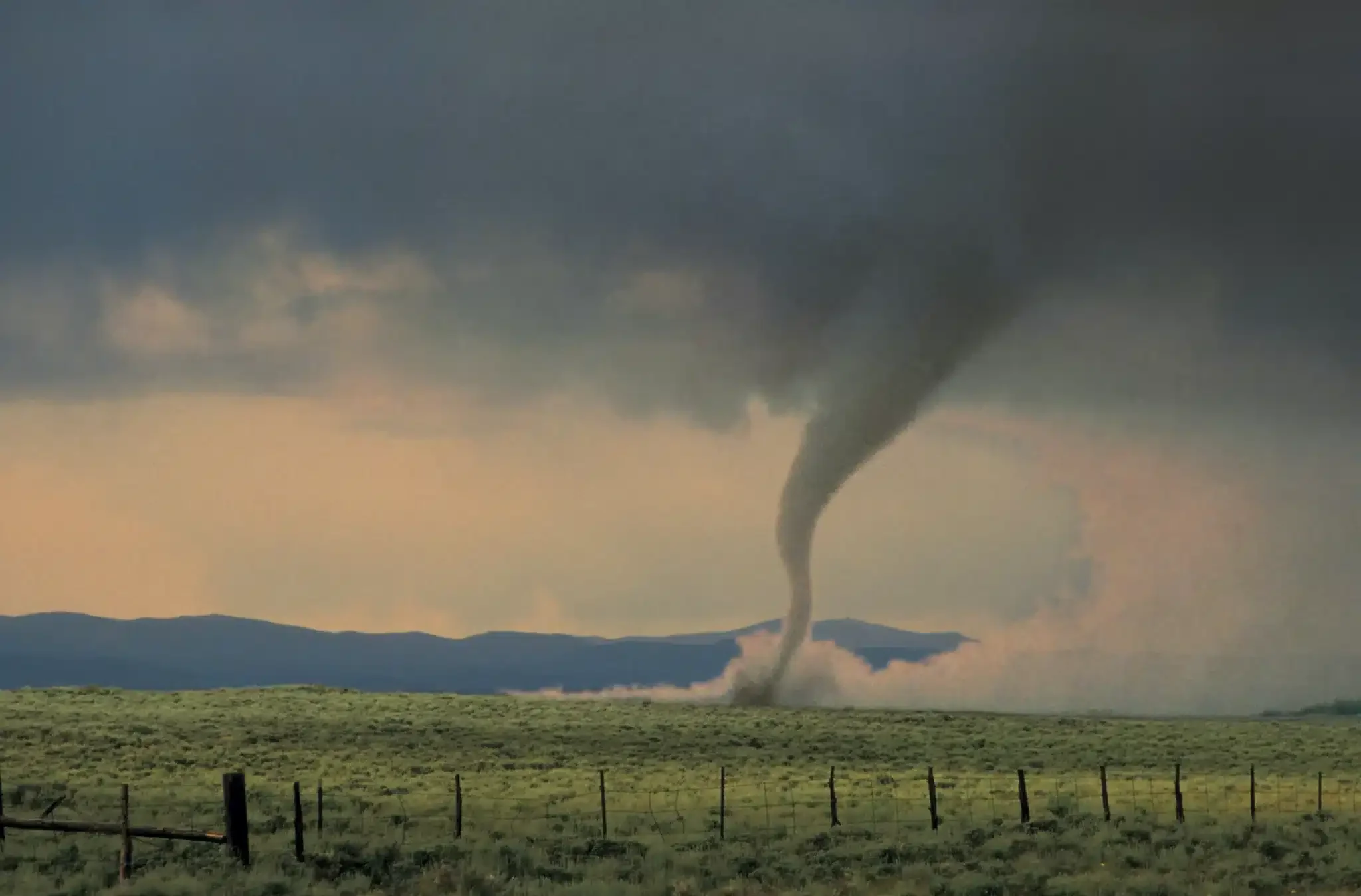 A tornado touches down in a rural landscape with a grassy field and a fence in the foreground, against a dramatically overcast sky.