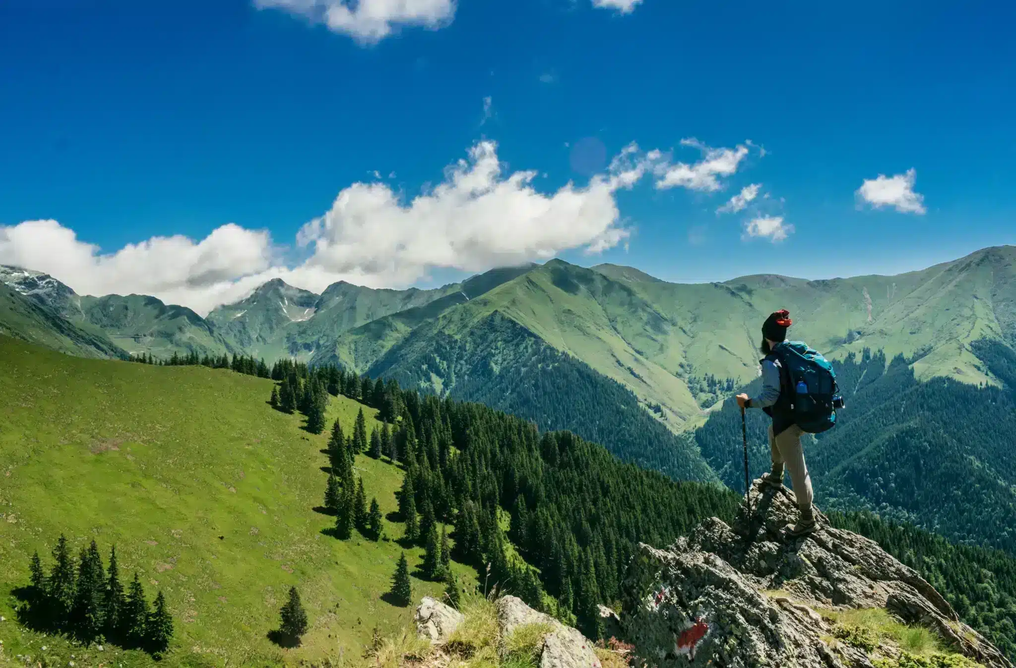 A hiker standing on a rocky outcrop, overlooking a panoramic view of lush green mountains with patches of snow under a clear blue sky with scattered clouds.