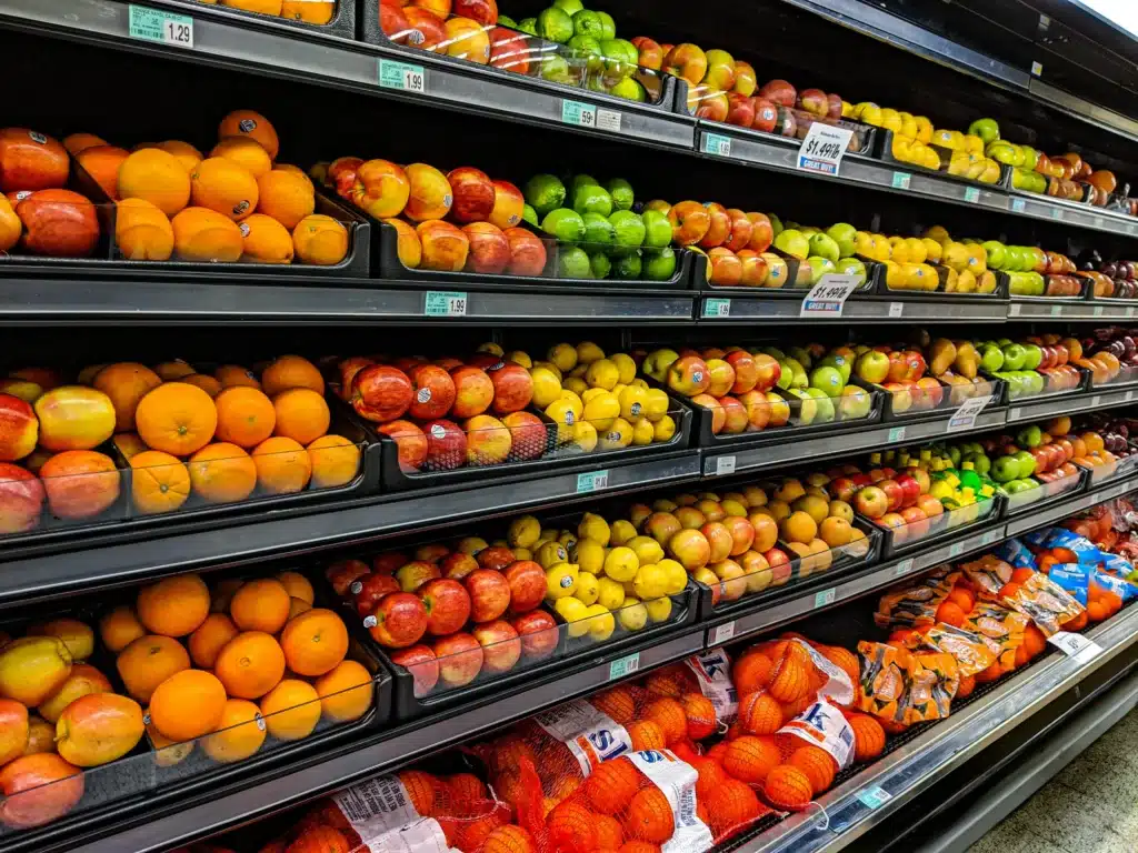 A colorful display of various fruits organized on supermarket shelves, including oranges, apples, and limes, with price tags visible above each selection.